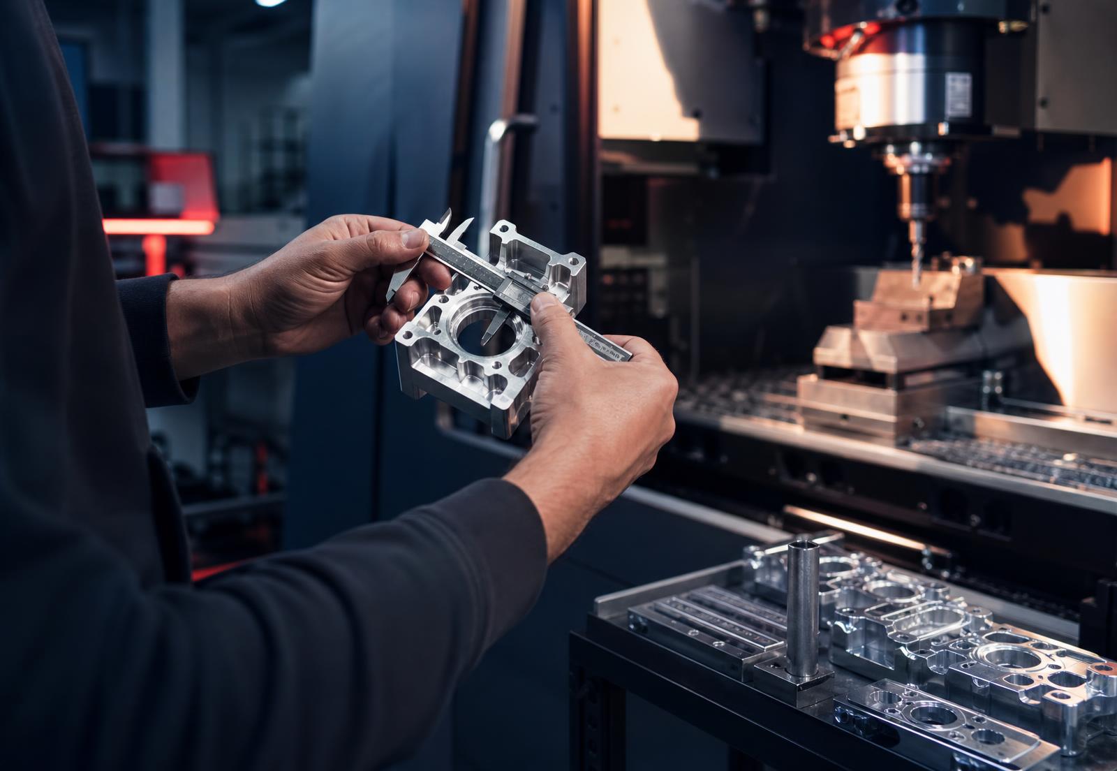 Machinist measuring a milled aluminum part beside a running CNC machining center