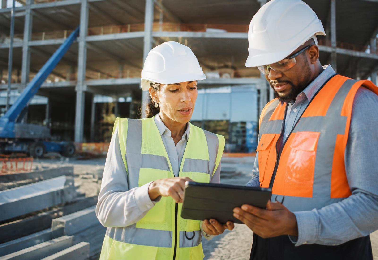 A woman contractor in a hard hat and safety vest reviewing digital plans on a tablet with a colleague at a commercial jobsite